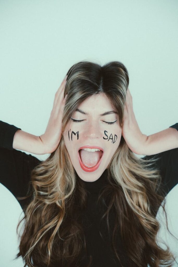 Young woman expresses sadness with words written on her face, hands over ears.